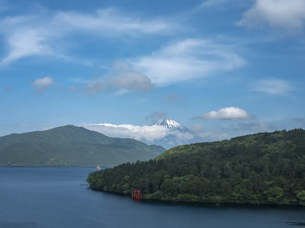 Snow-capped Mount Fuji rising above Lake Ashi, with the red torii gate of Hakone Shrine on the wooded shore