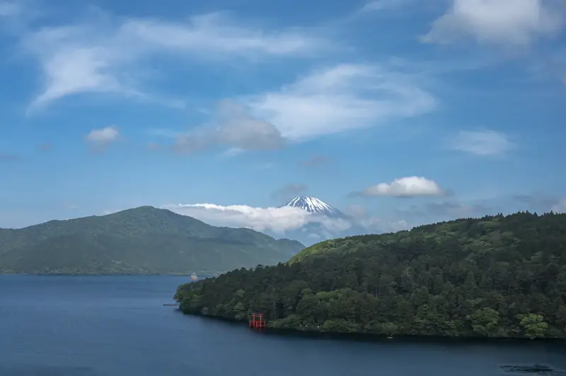 Snow-capped Mount Fuji rising above Lake Ashi, with the red torii gate of Hakone Shrine on the wooded shore