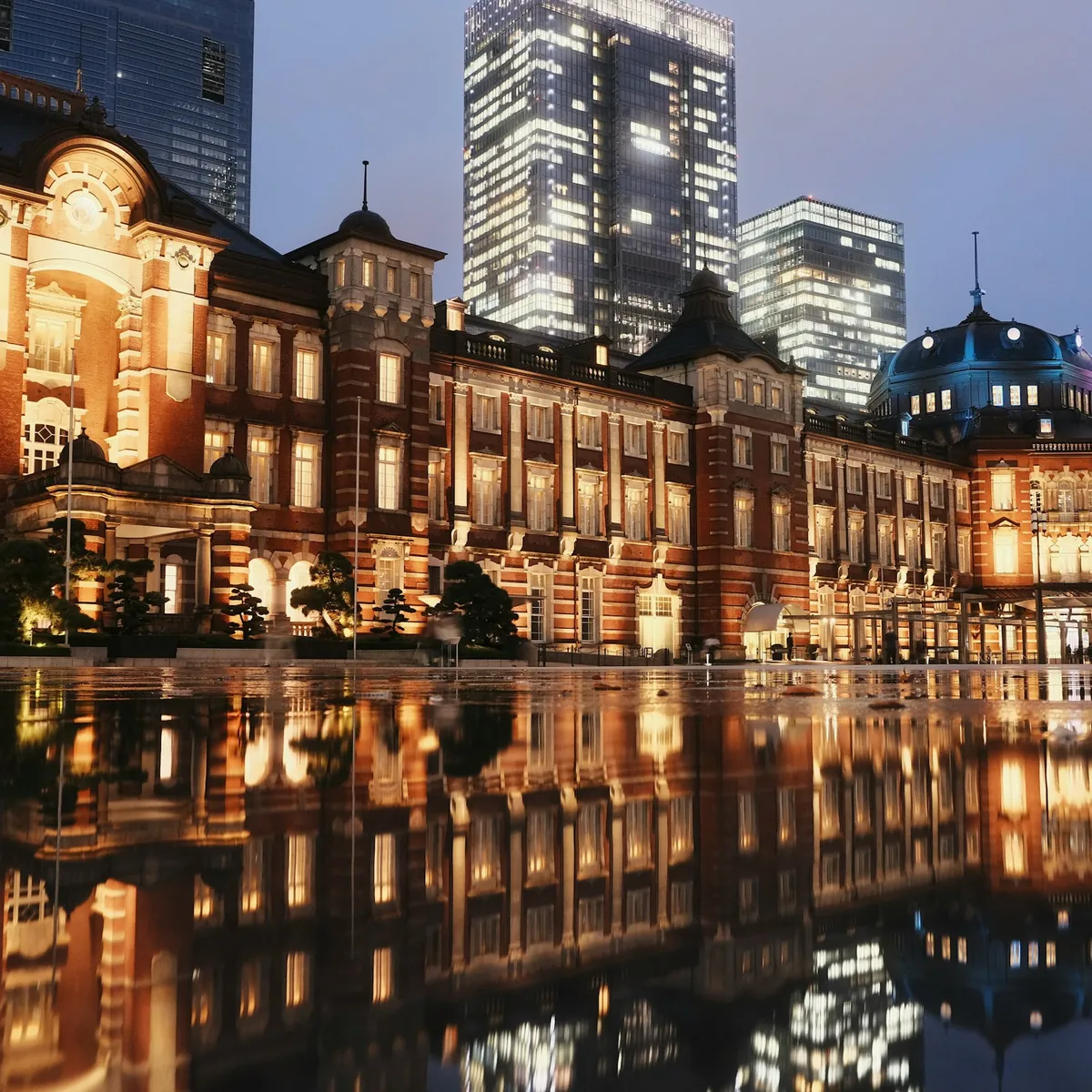 Tokyo Station red-brick Marunouchi facade lit at dusk and reflected in the wet plaza, with the towers of Marunouchi behind