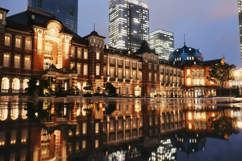 Tokyo Station red-brick Marunouchi facade lit at dusk and reflected in the wet plaza, with the towers of Marunouchi behind