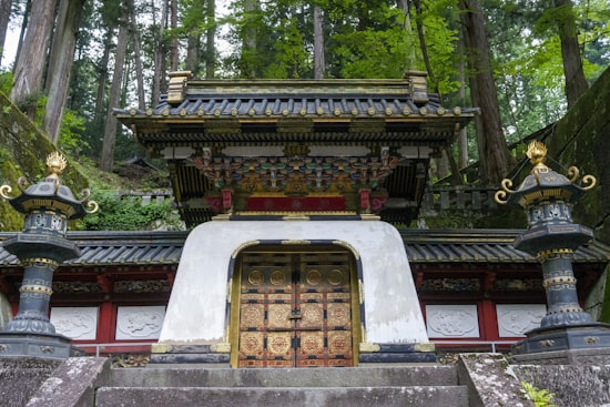 A gilded mausoleum gate flanked by bronze lanterns amid the giant cedars of Nikko